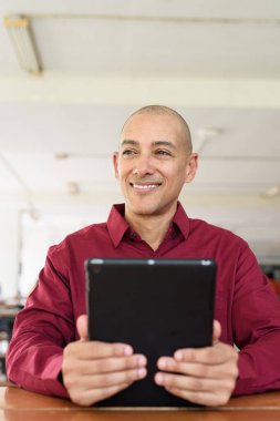 Burgundy renkli tişört giyen kendine güvenen bir adam devlet üniversitesinde ya da iş alanında kahve masasında tablet bilgisayar kullanıyor. Masadaki kahve fincanı sıradan bir hayat tarzı katıyor..