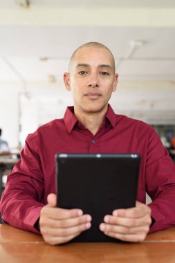 Burgundy renkli tişört giyen kendine güvenen bir adam devlet üniversitesinde ya da iş alanında kahve masasında tablet bilgisayar kullanıyor. Masadaki kahve fincanı sıradan bir hayat tarzı katıyor..