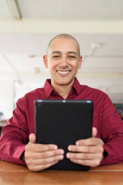 Burgundy renkli tişört giyen kendine güvenen bir adam devlet üniversitesinde ya da iş alanında kahve masasında tablet bilgisayar kullanıyor. Masadaki kahve fincanı sıradan bir hayat tarzı katıyor..