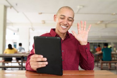 Burgundy renkli tişört giyen kendine güvenen bir adam devlet üniversitesinde ya da iş alanında kahve masasında tablet bilgisayar kullanıyor. Masadaki kahve fincanı sıradan bir hayat tarzı katıyor..