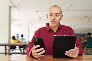 Burgundy renkli tişört giyen kendine güvenen bir adam devlet üniversitesinde ya da iş alanında kahve masasında tablet bilgisayar kullanıyor. Masadaki kahve fincanı sıradan bir hayat tarzı katıyor..