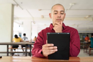 Burgundy renkli tişört giyen kendine güvenen bir adam devlet üniversitesinde ya da iş alanında kahve masasında tablet bilgisayar kullanıyor. Masadaki kahve fincanı sıradan bir hayat tarzı katıyor..