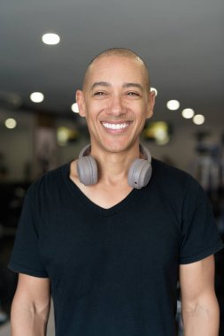 Spor salonunda antrenman yapan kel, İspanyol bir adam. Kaslı erkek, içeride özgüvenle egzersiz yapıyor, siyah gömlek ve kulaklık takıyor. Sağlıklı yaşam tarzı, güç ve sağlığa odaklanma..