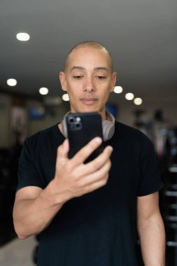 Spor salonunda antrenman yapan kel, İspanyol bir adam. Kaslı erkek, içeride özgüvenle egzersiz yapıyor, siyah gömlek ve kulaklık takıyor. Sağlıklı yaşam tarzı, güç ve sağlığa odaklanma..