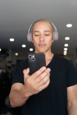 Spor salonunda antrenman yapan kel, İspanyol bir adam. Kaslı erkek, içeride özgüvenle egzersiz yapıyor, siyah gömlek ve kulaklık takıyor. Sağlıklı yaşam tarzı, güç ve sağlığa odaklanma..