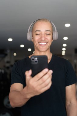 Spor salonunda antrenman yapan kel, İspanyol bir adam. Kaslı erkek, içeride özgüvenle egzersiz yapıyor, siyah gömlek ve kulaklık takıyor. Sağlıklı yaşam tarzı, güç ve sağlığa odaklanma..