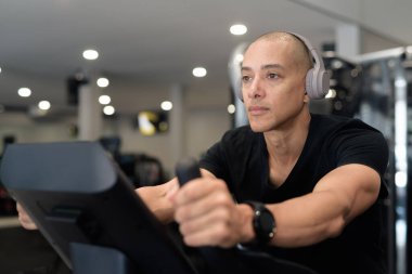 Spor salonunda antrenman yapan kel, İspanyol bir adam. Kaslı erkek, içeride özgüvenle egzersiz yapıyor, siyah gömlek ve kulaklık takıyor. Sağlıklı yaşam tarzı, güç ve sağlığa odaklanma..