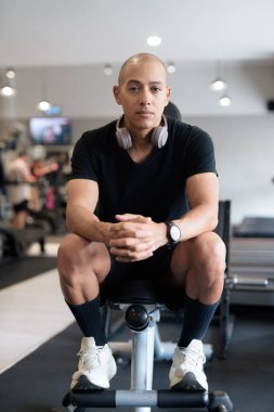 Spor salonunda antrenman yapan kel, İspanyol bir adam. Kaslı erkek, içeride özgüvenle egzersiz yapıyor, siyah gömlek ve kulaklık takıyor. Sağlıklı yaşam tarzı, güç ve sağlığa odaklanma..