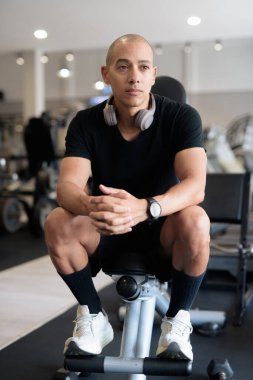 Spor salonunda antrenman yapan kel, İspanyol bir adam. Kaslı erkek, içeride özgüvenle egzersiz yapıyor, siyah gömlek ve kulaklık takıyor. Sağlıklı yaşam tarzı, güç ve sağlığa odaklanma..