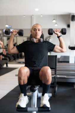 Spor salonunda antrenman yapan kel, İspanyol bir adam. Kaslı erkek, içeride özgüvenle egzersiz yapıyor, siyah gömlek ve kulaklık takıyor. Sağlıklı yaşam tarzı, güç ve sağlığa odaklanma..