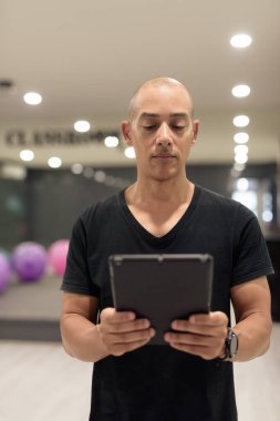 Spor salonunda antrenman yapan kel, İspanyol bir adam. Kaslı erkek, dijital tablet bilgisayar kullanıyor, kendine güveni var, siyah gömlek giyiyor. Sağlıklı yaşam tarzı, güç ve sağlığa odaklanma..