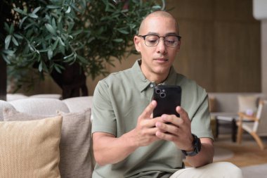 Gözlüklü Latin yetişkin adam otel lobisinde ya da bar salonunda otururken telefon kullanıyor. Modern dijital yaşam tarzı, günlük iş ortamı, seyahat ortamında iletişim ve bağlantı.