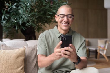 Gözlüklü Latin yetişkin adam otel lobisinde ya da bar salonunda otururken telefon kullanıyor. Modern dijital yaşam tarzı, günlük iş ortamı, seyahat ortamında iletişim ve bağlantı.