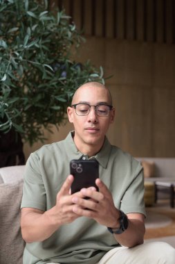 Gözlüklü Latin yetişkin adam otel lobisinde ya da bar salonunda otururken telefon kullanıyor. Modern dijital yaşam tarzı, günlük iş ortamı, seyahat ortamında iletişim ve bağlantı.