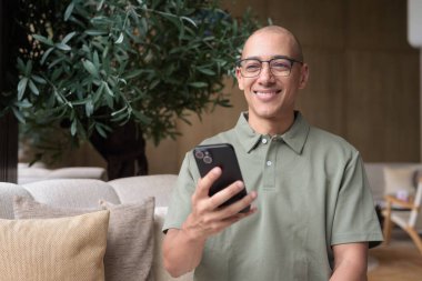 Gözlüklü Latin yetişkin adam otel lobisinde ya da bar salonunda otururken telefon kullanıyor. Modern dijital yaşam tarzı, günlük iş ortamı, seyahat ortamında iletişim ve bağlantı.