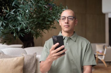 Gözlüklü Latin yetişkin adam otel lobisinde ya da bar salonunda otururken telefon kullanıyor. Modern dijital yaşam tarzı, günlük iş ortamı, seyahat ortamında iletişim ve bağlantı.