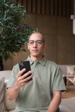 Gözlüklü Latin yetişkin adam otel lobisinde ya da bar salonunda otururken telefon kullanıyor. Modern dijital yaşam tarzı, günlük iş ortamı, seyahat ortamında iletişim ve bağlantı.