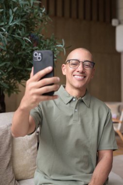 Gözlüklü Latin yetişkin adam otel lobisinde ya da bar salonunda otururken telefon kullanıyor. Modern dijital yaşam tarzı, günlük iş ortamı, seyahat ortamında iletişim ve bağlantı.