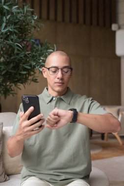 Gözlüklü Latin yetişkin adam otel lobisinde ya da bar salonunda otururken telefon kullanıyor. Modern dijital yaşam tarzı, günlük iş ortamı, seyahat ortamında iletişim ve bağlantı.