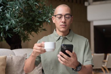 Gözlüklü Latin yetişkin adam otel lobisinde ya da bar salonunda otururken telefon kullanıyor. Modern dijital yaşam tarzı, günlük iş ortamı, seyahat ortamında iletişim ve bağlantı.