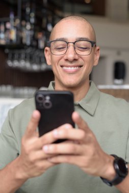 Gözlüklü Latin yetişkin adam otel lobisinde ya da bar salonunda otururken telefon kullanıyor. Modern dijital yaşam tarzı, günlük iş ortamı, seyahat ortamında iletişim ve bağlantı.