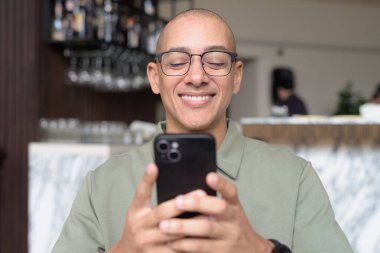 Gözlüklü Latin yetişkin adam otel lobisinde ya da bar salonunda otururken telefon kullanıyor. Modern dijital yaşam tarzı, günlük iş ortamı, seyahat ortamında iletişim ve bağlantı.
