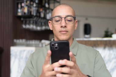 Gözlüklü Latin yetişkin adam otel lobisinde ya da bar salonunda otururken telefon kullanıyor. Modern dijital yaşam tarzı, günlük iş ortamı, seyahat ortamında iletişim ve bağlantı.