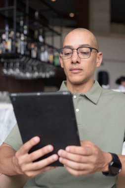 Gözlüklü Latin yetişkin adam otel lobisinde ya da bar salonunda otururken telefon kullanıyor. Modern dijital yaşam tarzı, günlük iş ortamı, seyahat ortamında iletişim ve bağlantı.