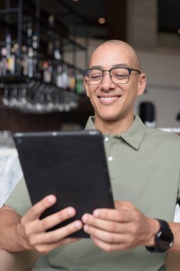 Gözlüklü Latin yetişkin adam otel lobisinde ya da bar salonunda otururken telefon kullanıyor. Modern dijital yaşam tarzı, günlük iş ortamı, seyahat ortamında iletişim ve bağlantı.