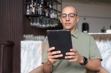 Gözlüklü Latin yetişkin adam otel lobisinde ya da bar salonunda otururken telefon kullanıyor. Modern dijital yaşam tarzı, günlük iş ortamı, seyahat ortamında iletişim ve bağlantı.