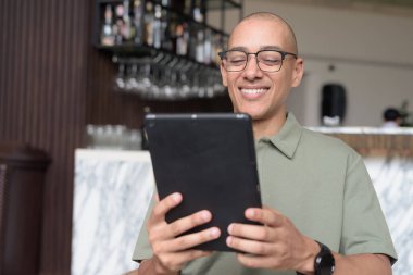 Gözlüklü Latin yetişkin adam otel lobisinde ya da bar salonunda otururken telefon kullanıyor. Modern dijital yaşam tarzı, günlük iş ortamı, seyahat ortamında iletişim ve bağlantı.