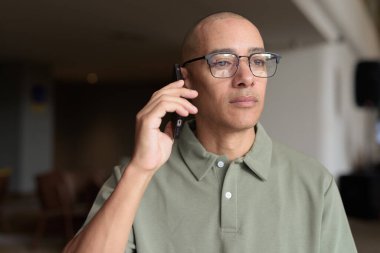 Gözlüklü Latin yetişkin adam otel lobisinde ya da bar salonunda otururken telefon kullanıyor. Modern dijital yaşam tarzı, günlük iş ortamı, seyahat ortamında iletişim ve bağlantı.