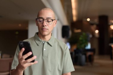 Gözlüklü Latin yetişkin adam otel lobisinde ya da bar salonunda otururken telefon kullanıyor. Modern dijital yaşam tarzı, günlük iş ortamı, seyahat ortamında iletişim ve bağlantı.