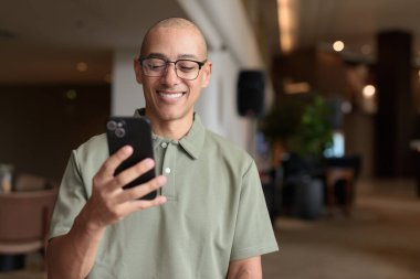 Gözlüklü Latin yetişkin adam otel lobisinde ya da bar salonunda otururken telefon kullanıyor. Modern dijital yaşam tarzı, günlük iş ortamı, seyahat ortamında iletişim ve bağlantı.