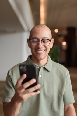 Gözlüklü Latin yetişkin adam otel lobisinde ya da bar salonunda otururken telefon kullanıyor. Modern dijital yaşam tarzı, günlük iş ortamı, seyahat ortamında iletişim ve bağlantı.