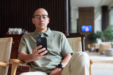 Gözlüklü Latin yetişkin adam otel lobisinde ya da bar salonunda otururken telefon kullanıyor. Modern dijital yaşam tarzı, günlük iş ortamı, seyahat ortamında iletişim ve bağlantı.