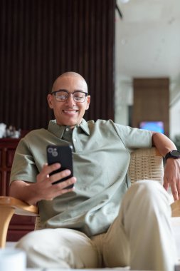 Gözlüklü Latin yetişkin adam otel lobisinde ya da bar salonunda otururken telefon kullanıyor. Modern dijital yaşam tarzı, günlük iş ortamı, seyahat ortamında iletişim ve bağlantı.