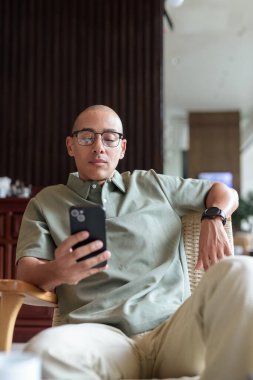 Gözlüklü Latin yetişkin adam otel lobisinde ya da bar salonunda otururken telefon kullanıyor. Modern dijital yaşam tarzı, günlük iş ortamı, seyahat ortamında iletişim ve bağlantı.