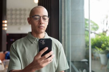 Gözlüklü Latin yetişkin adam otel lobisinde ya da bar salonunda otururken telefon kullanıyor. Modern dijital yaşam tarzı, günlük iş ortamı, seyahat ortamında iletişim ve bağlantı.