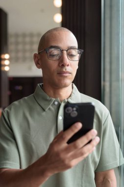 Gözlüklü Latin yetişkin adam otel lobisinde ya da bar salonunda otururken telefon kullanıyor. Modern dijital yaşam tarzı, günlük iş ortamı, seyahat ortamında iletişim ve bağlantı.