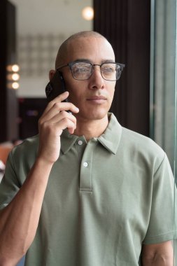 Gözlüklü Latin yetişkin adam otel lobisinde ya da bar salonunda otururken telefon kullanıyor. Modern dijital yaşam tarzı, günlük iş ortamı, seyahat ortamında iletişim ve bağlantı.