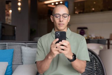 Gözlüklü Latin yetişkin adam otel lobisinde ya da bar salonunda otururken telefon kullanıyor. Modern dijital yaşam tarzı, günlük iş ortamı, seyahat ortamında iletişim ve bağlantı.