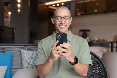 Gözlüklü Latin yetişkin adam otel lobisinde ya da bar salonunda otururken telefon kullanıyor. Modern dijital yaşam tarzı, günlük iş ortamı, seyahat ortamında iletişim ve bağlantı.