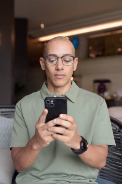 Gözlüklü Latin yetişkin adam otel lobisinde ya da bar salonunda otururken telefon kullanıyor. Modern dijital yaşam tarzı, günlük iş ortamı, seyahat ortamında iletişim ve bağlantı.