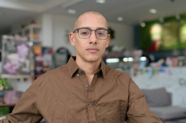 40 'lı yaşlarda kel bir adam kafeteryada oturuyor, günlük gömlek ve gözlük takıyor. Kendinden emin ve mutlu bir ifade. Kentsel yaşam tarzı, modern adam, rahat ortamda yetişkin portresi.