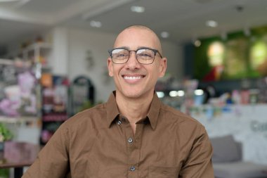 40 'lı yaşlarda kel bir adam kafeteryada oturuyor, günlük gömlek ve gözlük takıyor. Kendinden emin ve mutlu bir ifade. Kentsel yaşam tarzı, modern adam, rahat ortamda yetişkin portresi.