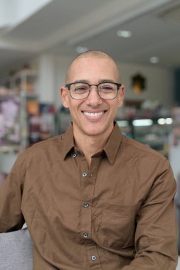 40 'lı yaşlarda kel bir adam kafeteryada oturuyor, günlük gömlek ve gözlük takıyor. Kendinden emin ve mutlu bir ifade. Kentsel yaşam tarzı, modern adam, rahat ortamda yetişkin portresi.