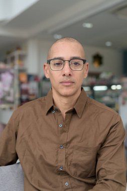 40 'lı yaşlarda kel bir adam kafeteryada oturuyor, günlük gömlek ve gözlük takıyor. Kendinden emin ve mutlu bir ifade. Kentsel yaşam tarzı, modern adam, rahat ortamda yetişkin portresi.