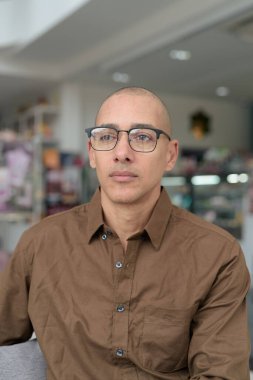 40 'lı yaşlarda kel bir adam kafeteryada oturuyor, günlük gömlek ve gözlük takıyor. Kendinden emin ve mutlu bir ifade. Kentsel yaşam tarzı, modern adam, rahat ortamda yetişkin portresi.