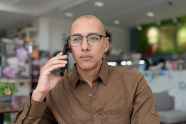 Gözlüklü kel Latin adam kafeteryada akıllı telefon kullanarak oturuyor. Sıradan bir gömlek giyiyor, gülümsüyor ve kendinden emin. Modern dijital yaşam tarzı, iletişim, mobil teknoloji, kentsel kahve dükkanı sahnesi.